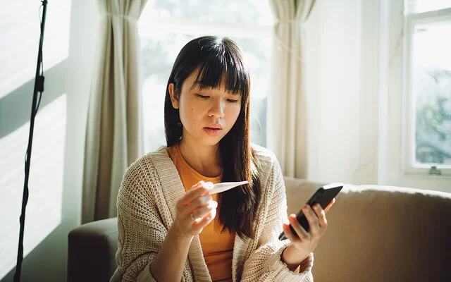 Eine junge Frau blickt auf ein Fieberthermometer in ihrer Hand. In der anderen Hand hält sie ein Smartphone.